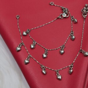 Two 925 sterling silver anklets featuring evenly spaced, smooth heart drop charms on a delicate chain, arranged in a coiled pattern on a textured red leather surface.