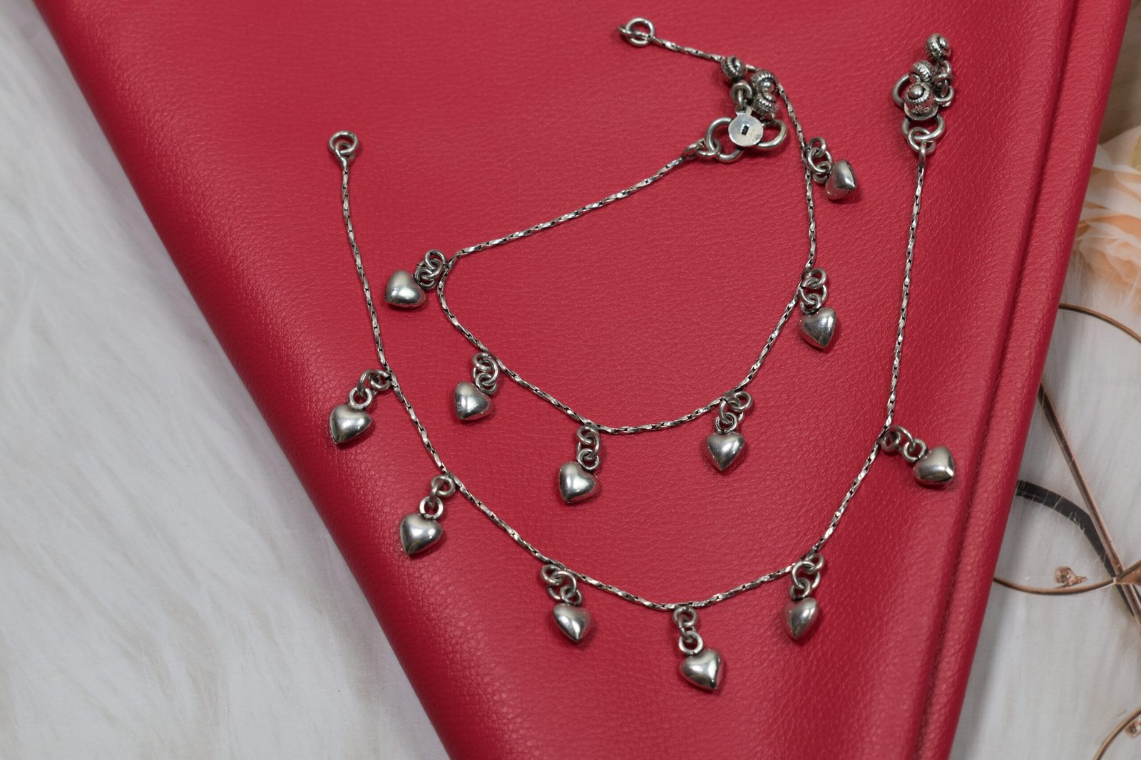 desireable-chained-heart-drop-silver-anklets2 Two 925 sterling silver anklets featuring evenly spaced, smooth heart drop charms on a delicate chain, arranged in a coiled pattern on a textured red leather surface.