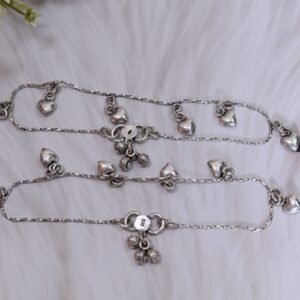A pair of silver anklets with dangling heart charms resting on a plush white faux fur surface, featuring an S-hook clasp stamped with '925' alongside small ghungroo bells and vintage glasses.