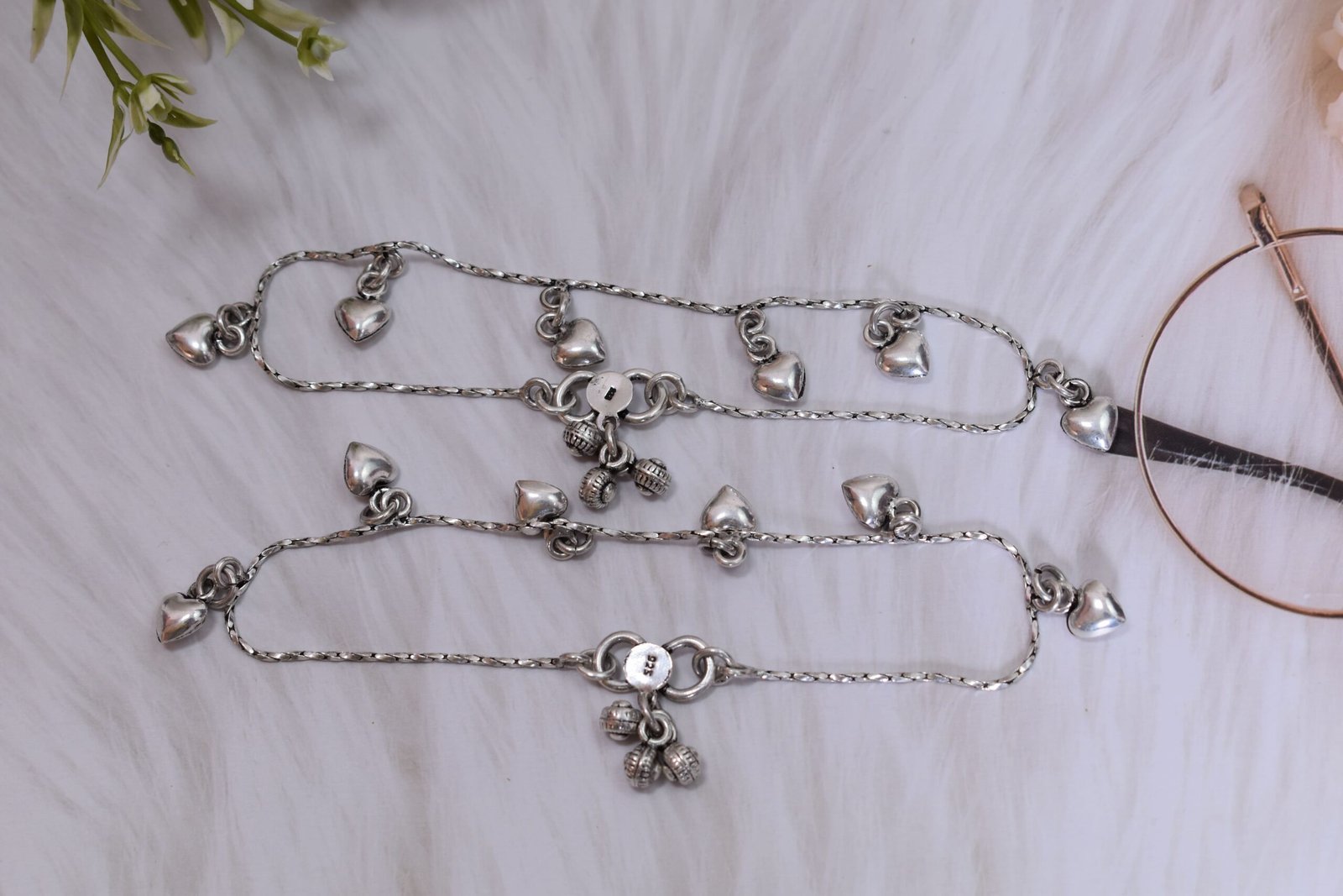 desireable-chained-heart-drop-silver-anklets3 A pair of silver anklets with dangling heart charms resting on a plush white faux fur surface, featuring an S-hook clasp stamped with '925' alongside small ghungroo bells and vintage glasses.