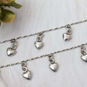 Macro close-up of two parallel silver anklet chains resting on a white textured surface, focusing on the shiny, puffed heart drop charms suspended from small rings.