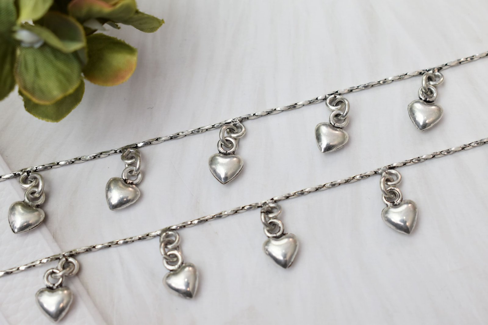 desireable-chained-heart-drop-silver-anklets4 Macro close-up of two parallel silver anklet chains resting on a white textured surface, focusing on the shiny, puffed heart drop charms suspended from small rings.