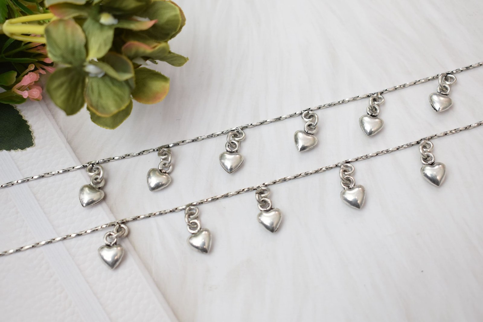 desireable-chained-heart-drop-silver-anklets5 Detailed diagonal view of two silver chains featuring dangling heart-shaped beads, highlighting the twisted texture of the main chain against a soft white background.
