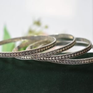 Four thin 925 sterling silver bangles overlapping on a soft white surface, displaying a continuous sequence of raised heart motifs framed by a textured, oxidized border.