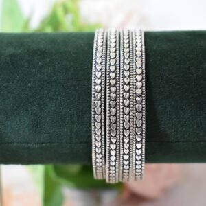 Four thin 925 sterling silver bangles overlapping on a soft white surface, displaying a continuous sequence of raised heart motifs framed by a textured, oxidized border.