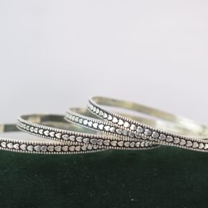 Four thin 925 sterling silver bangles overlapping on a soft white surface, displaying a continuous sequence of raised heart motifs framed by a textured, oxidized border.