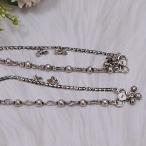 a pair of 925 sterling silver anklets resting on a white textured surface and soft faux fur, showcasing a fusion design of smooth silver balls and a linked chain with small dangling charms, finishing with ghungroo clasps near floral accents.