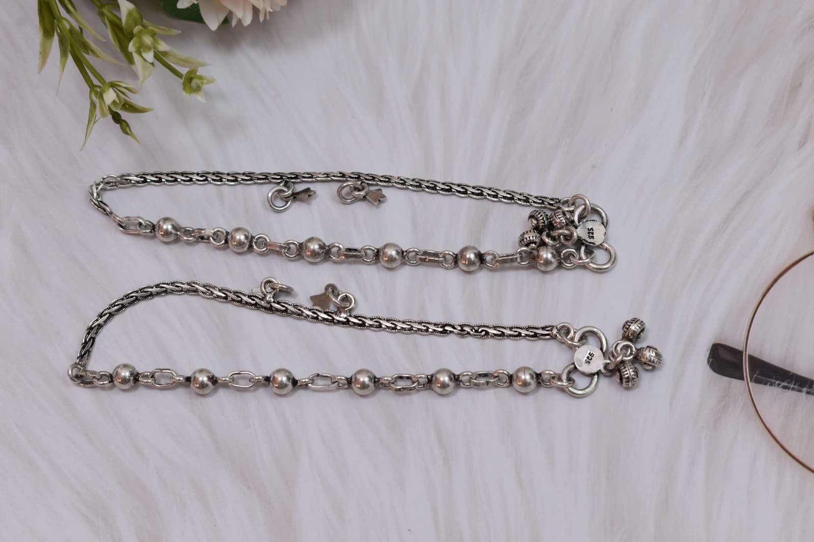 impressive-ball-fusion-chained-silver-anklets1 a pair of 925 sterling silver anklets resting on a white textured surface and soft faux fur, showcasing a fusion design of smooth silver balls and a linked chain with small dangling charms, finishing with ghungroo clasps near floral accents.