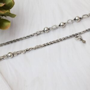 a pair of 925 sterling silver anklets resting on a white textured surface and soft faux fur, showcasing a fusion design of smooth silver balls and a linked chain with small dangling charms, finishing with ghungroo clasps near floral accents.