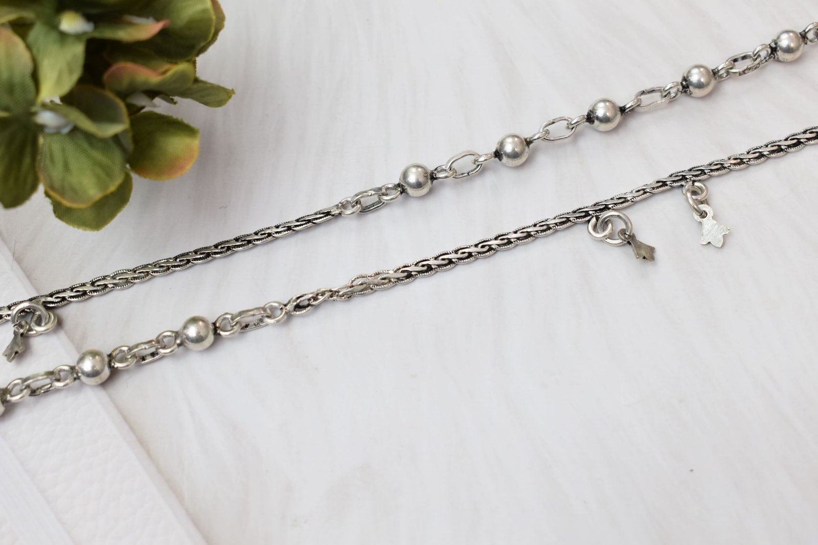 impressive-ball-fusion-chained-silver-anklets2 a pair of 925 sterling silver anklets resting on a white textured surface and soft faux fur, showcasing a fusion design of smooth silver balls and a linked chain with small dangling charms, finishing with ghungroo clasps near floral accents.
