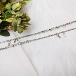 a pair of 925 sterling silver anklets resting on a white textured surface and soft faux fur, showcasing a fusion design of smooth silver balls and a linked chain with small dangling charms, finishing with ghungroo clasps near floral accents.
