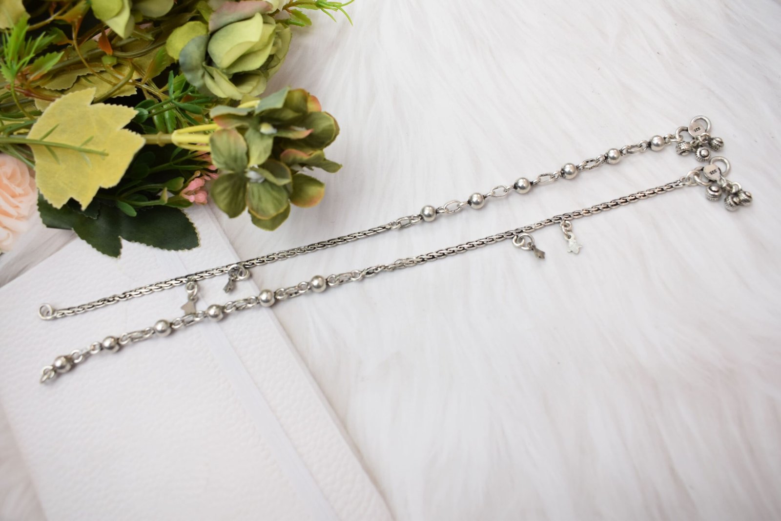impressive-ball-fusion-chained-silver-anklets3 a pair of 925 sterling silver anklets resting on a white textured surface and soft faux fur, showcasing a fusion design of smooth silver balls and a linked chain with small dangling charms, finishing with ghungroo clasps near floral accents.