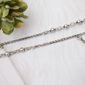 a pair of 925 sterling silver anklets resting on a white textured surface and soft faux fur, showcasing a fusion design of smooth silver balls and a linked chain with small dangling charms, finishing with ghungroo clasps near floral accents.