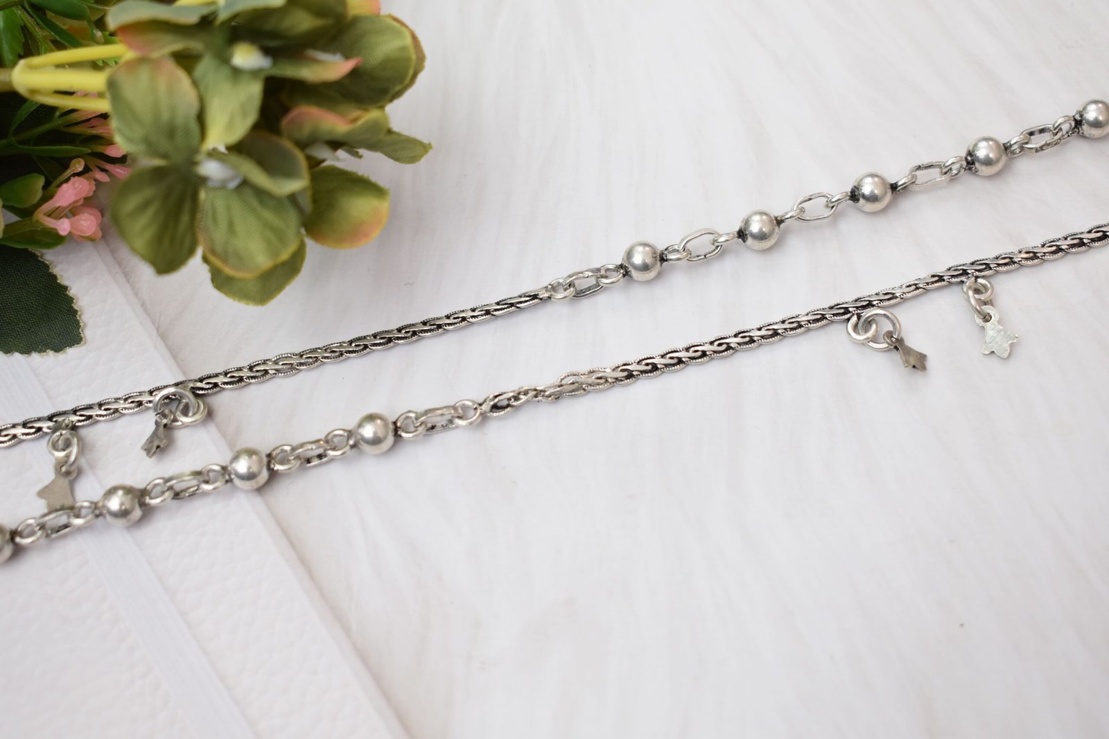 impressive-ball-fusion-chained-silver-anklets4 a pair of 925 sterling silver anklets resting on a white textured surface and soft faux fur, showcasing a fusion design of smooth silver balls and a linked chain with small dangling charms, finishing with ghungroo clasps near floral accents.