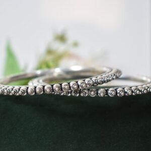 A pair of overlapping 925 sterling silver kadas resting on a soft white surface, showcasing a highly textured design that fuses larger polished silver balls with a smaller beaded edge against a dark, oxidized background.