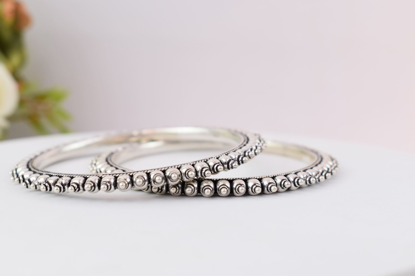 intricate-double-ball-fusion-everyday-wear-silver-kada3 A pair of overlapping 925 sterling silver kadas resting on a soft white surface, showcasing a highly textured design that fuses larger polished silver balls with a smaller beaded edge against a dark, oxidized background.