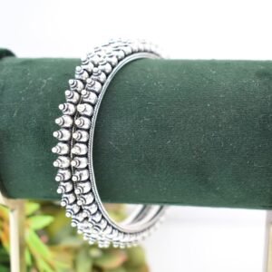A pair of overlapping 925 sterling silver kadas resting on a soft white surface, showcasing a highly textured design that fuses larger polished silver balls with a smaller beaded edge against a dark, oxidized background.