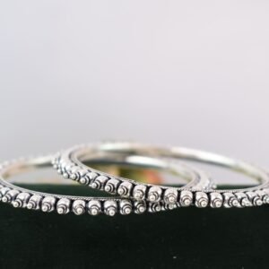 A pair of overlapping 925 sterling silver kadas resting on a soft white surface, showcasing a highly textured design that fuses larger polished silver balls with a smaller beaded edge against a dark, oxidized background.
