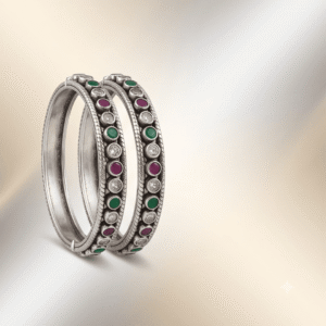 A pair of 925 sterling silver kadas standing upright against a soft gradient background, featuring a continuous sequence of round green, clear white, and ruby red cut stones bordered by textured silver detailing.