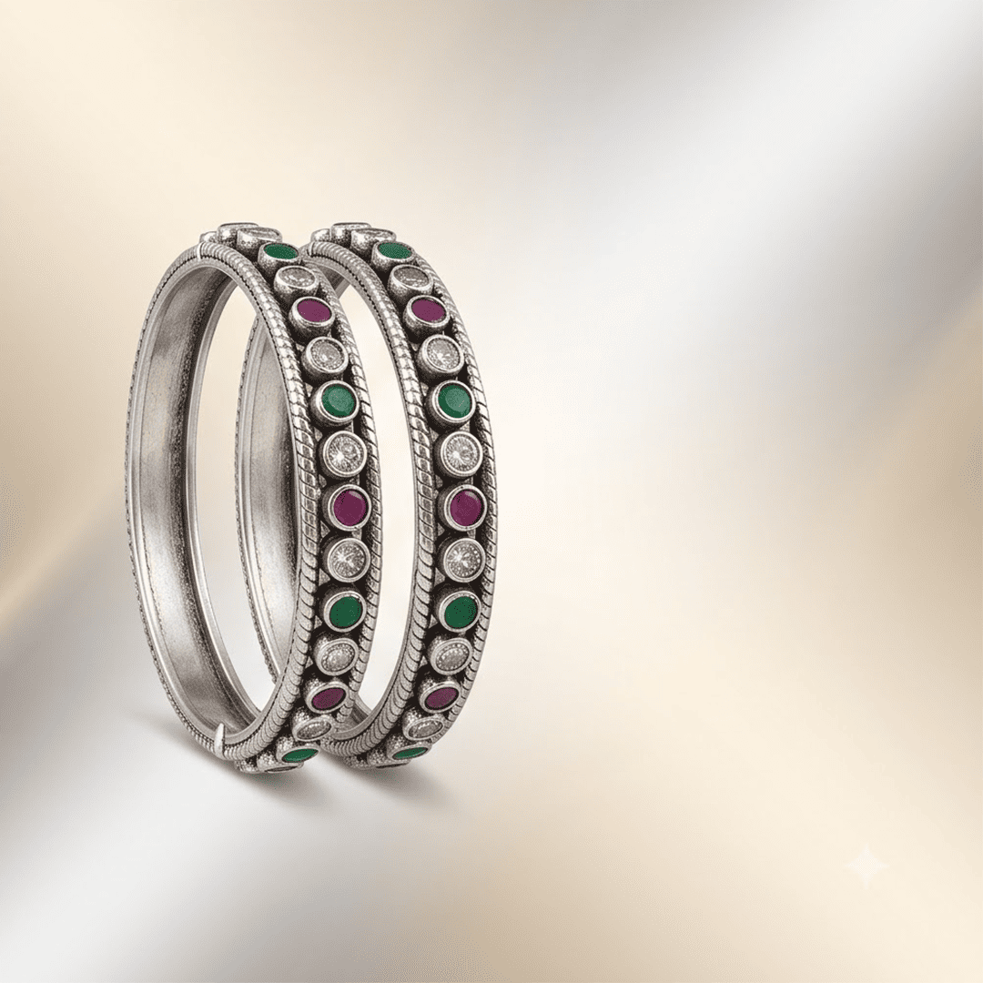 round-green-white-ruby-colour-cutstone-sequence-silver-kada.1 A pair of 925 sterling silver kadas standing upright against a soft gradient background, featuring a continuous sequence of round green, clear white, and ruby red cut stones bordered by textured silver detailing.