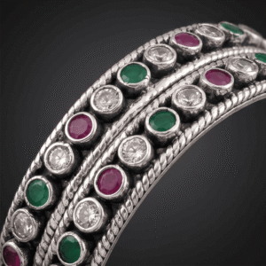 A pair of 925 sterling silver kadas standing upright against a soft gradient background, featuring a continuous sequence of round green, clear white, and ruby red cut stones bordered by textured silver detailing.