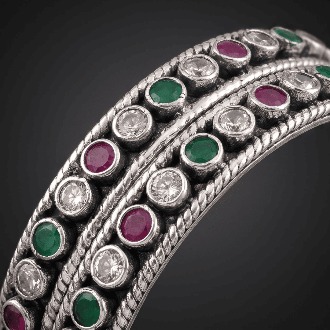round-green-white-ruby-colour-cutstone-sequence-silver-kada.2 A pair of 925 sterling silver kadas standing upright against a soft gradient background, featuring a continuous sequence of round green, clear white, and ruby red cut stones bordered by textured silver detailing.