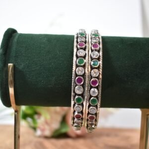 A pair of 925 sterling silver kadas standing upright against a soft gradient background, featuring a continuous sequence of round green, clear white, and ruby red cut stones bordered by textured silver detailing.