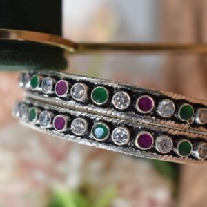 A pair of 925 sterling silver kadas standing upright against a soft gradient background, featuring a continuous sequence of round green, clear white, and ruby red cut stones bordered by textured silver detailing.