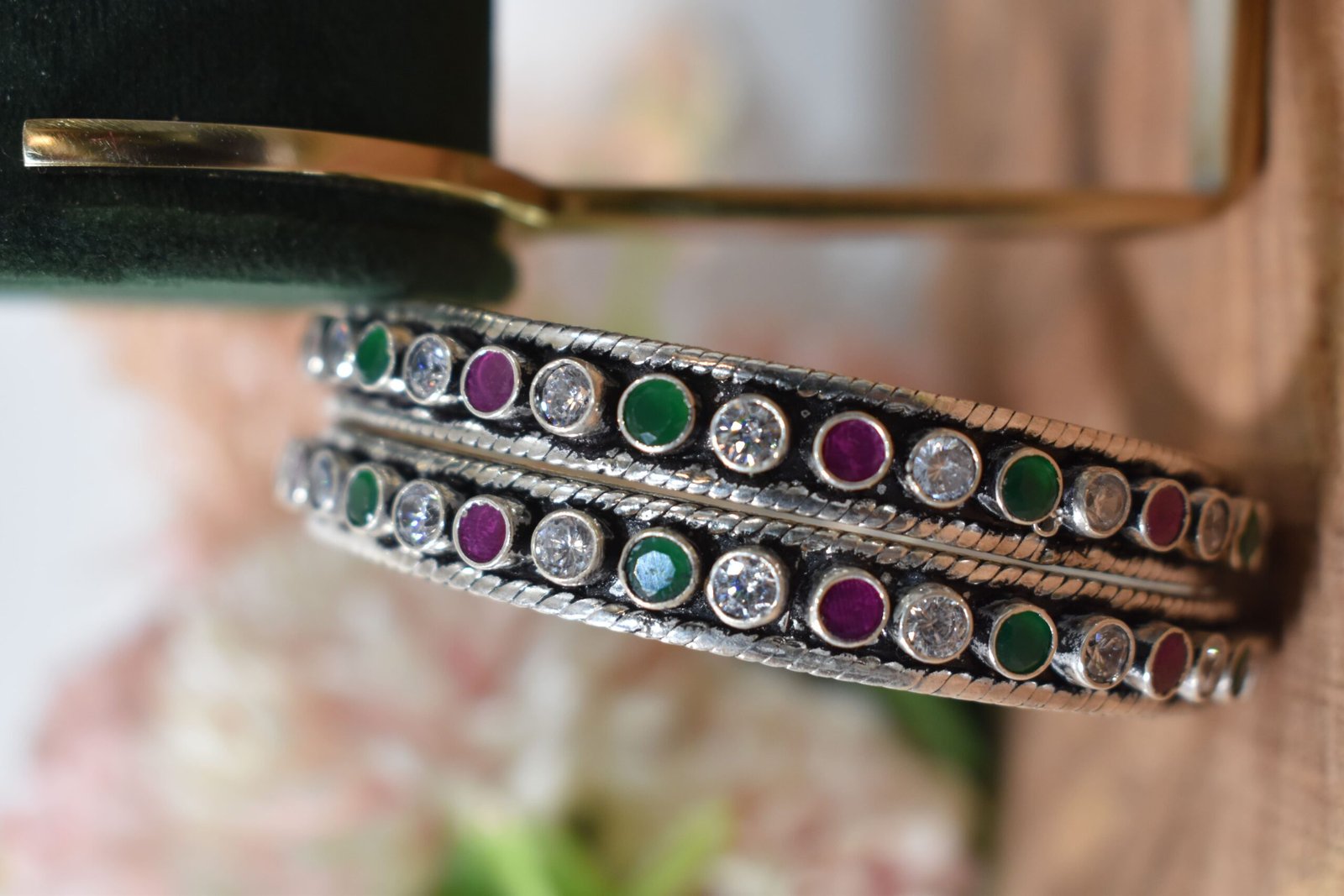 round-green-white-ruby-colour-cutstone-sequence-silver-kada.5 A pair of 925 sterling silver kadas standing upright against a soft gradient background, featuring a continuous sequence of round green, clear white, and ruby red cut stones bordered by textured silver detailing.