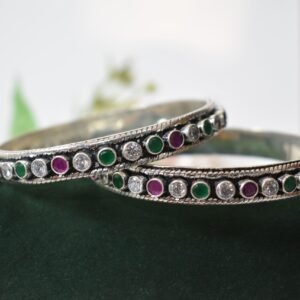 A pair of 925 sterling silver kadas standing upright against a soft gradient background, featuring a continuous sequence of round green, clear white, and ruby red cut stones bordered by textured silver detailing.