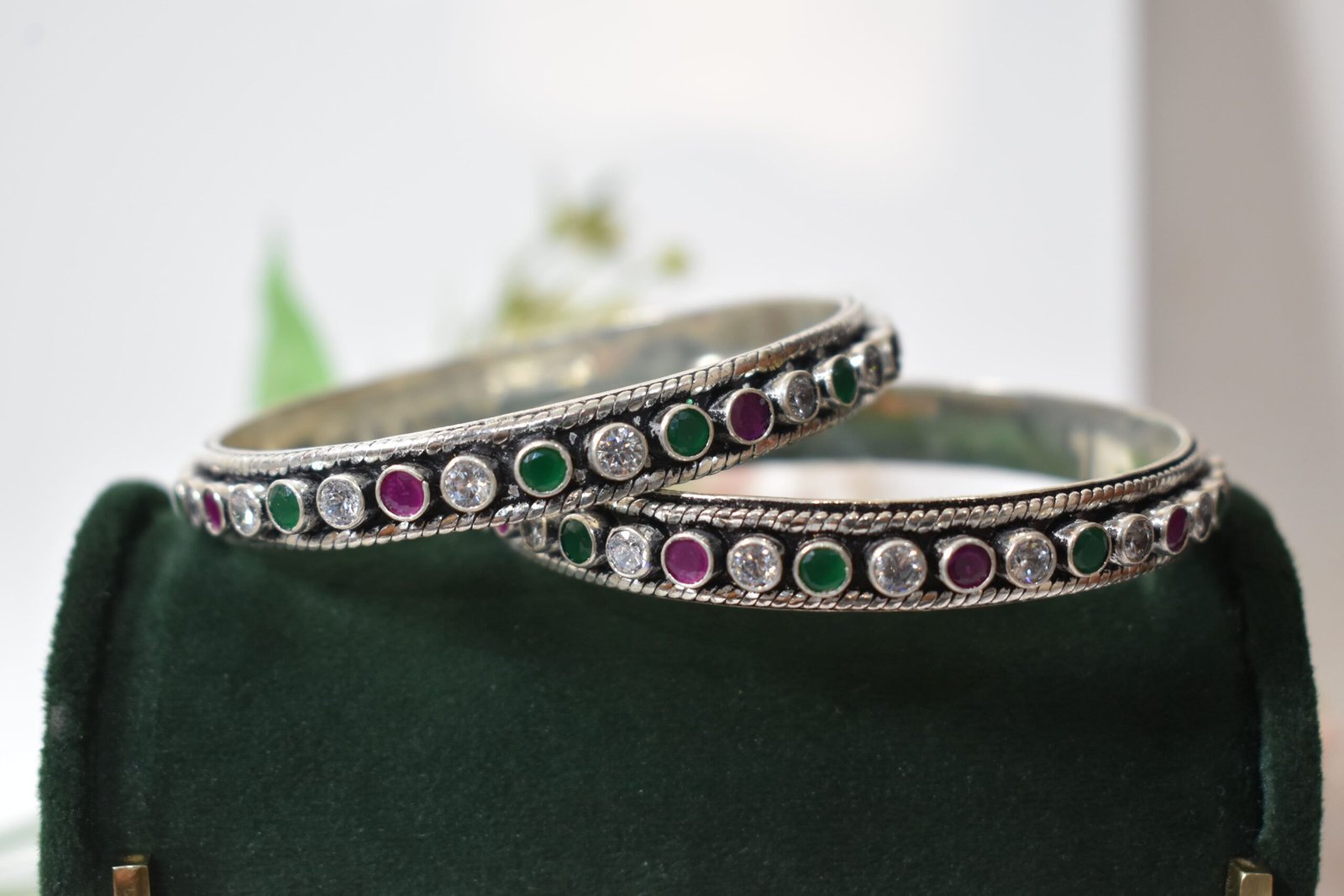 round-green-white-ruby-colour-cutstone-sequence-silver-kada.6 A pair of 925 sterling silver kadas standing upright against a soft gradient background, featuring a continuous sequence of round green, clear white, and ruby red cut stones bordered by textured silver detailing.