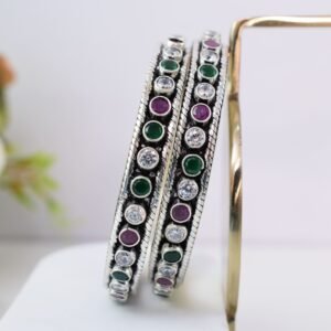 A pair of 925 sterling silver kadas standing upright against a soft gradient background, featuring a continuous sequence of round green, clear white, and ruby red cut stones bordered by textured silver detailing.