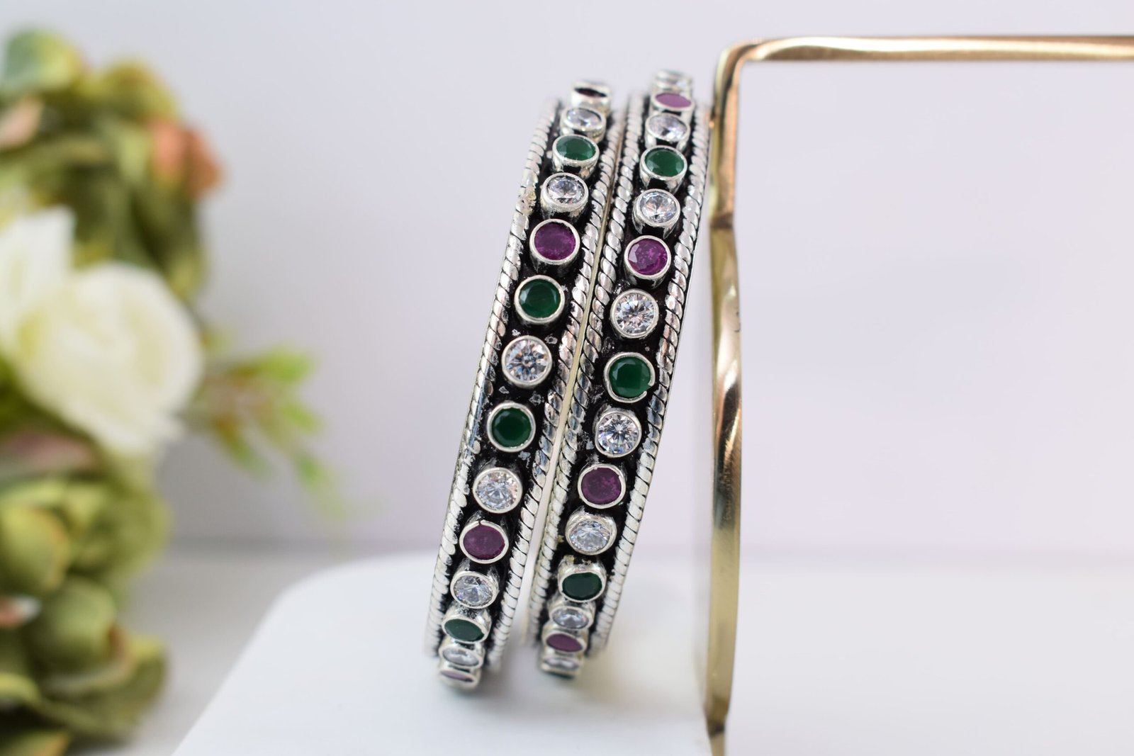 round-green-white-ruby-colour-cutstone-sequence-silver-kada.7 A pair of 925 sterling silver kadas standing upright against a soft gradient background, featuring a continuous sequence of round green, clear white, and ruby red cut stones bordered by textured silver detailing.