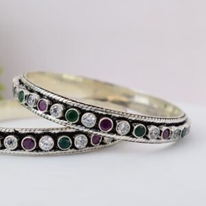 A pair of 925 sterling silver kadas standing upright against a soft gradient background, featuring a continuous sequence of round green, clear white, and ruby red cut stones bordered by textured silver detailing.