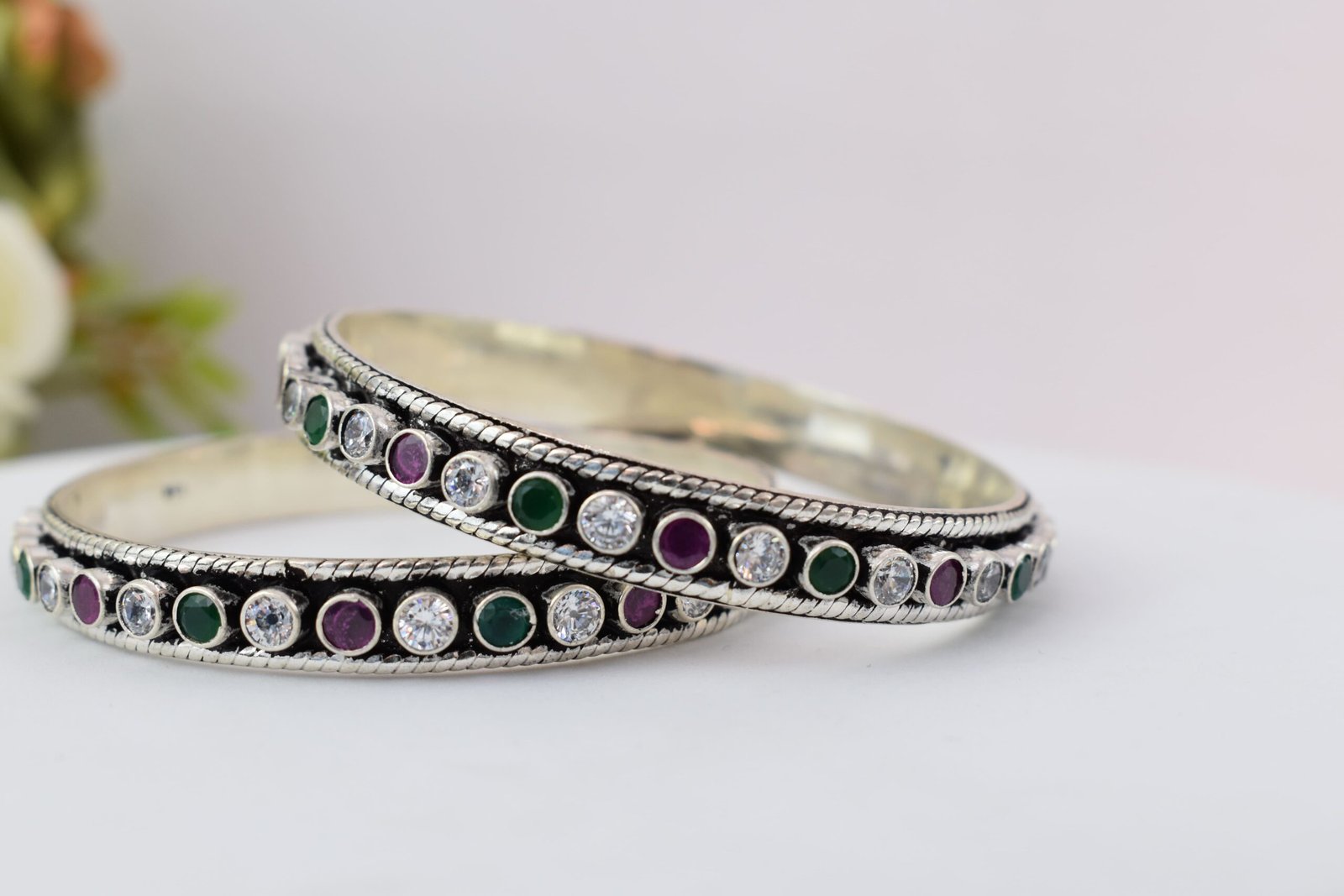 round-green-white-ruby-colour-cutstone-sequence-silver-kada.8 A pair of 925 sterling silver kadas standing upright against a soft gradient background, featuring a continuous sequence of round green, clear white, and ruby red cut stones bordered by textured silver detailing.