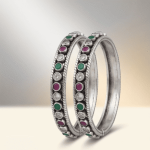 A pair of 925 sterling silver kadas standing upright against a soft gradient background, featuring a continuous sequence of round green, clear white, and ruby red cut stones bordered by textured silver detailing.