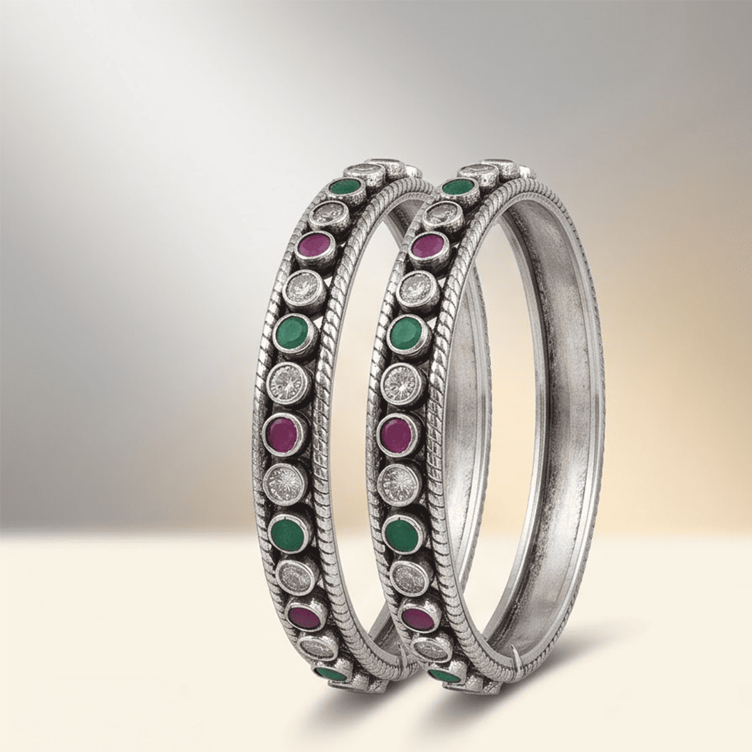 round-green-white-ruby-colour-cutstone-sequence-silver-kada.9 A pair of 925 sterling silver kadas standing upright against a soft gradient background, featuring a continuous sequence of round green, clear white, and ruby red cut stones bordered by textured silver detailing.