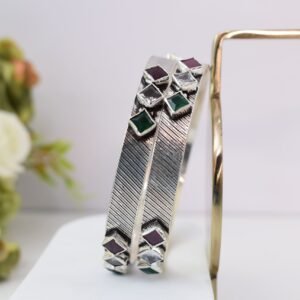 Close-up of two 925 sterling silver kadas standing upright, featuring a diagonally textured silver band and a sequence of square-cut green, white, and red stones set along the edges against a soft floral background.