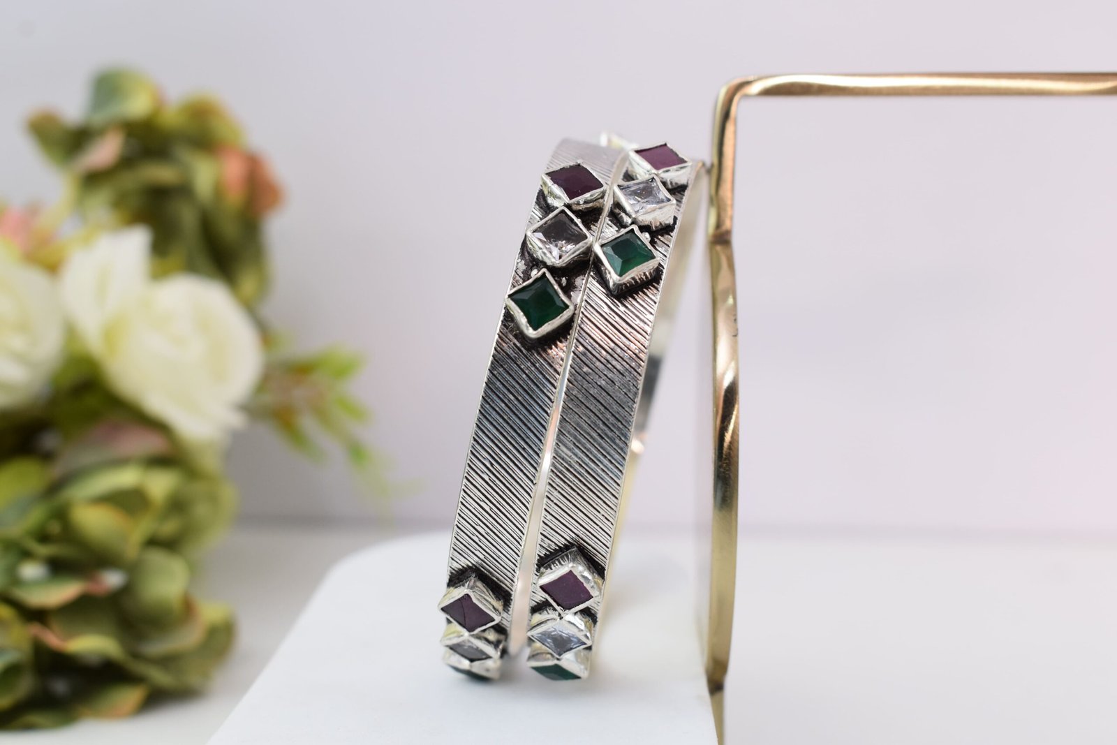 square-green-white-red-sequence-silver-kada1 Close-up of two 925 sterling silver kadas standing upright, featuring a diagonally textured silver band and a sequence of square-cut green, white, and red stones set along the edges against a soft floral background.