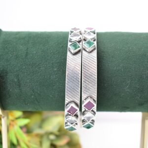 Close-up of two 925 sterling silver kadas standing upright, featuring a diagonally textured silver band and a sequence of square-cut green, white, and red stones set along the edges against a soft floral background.