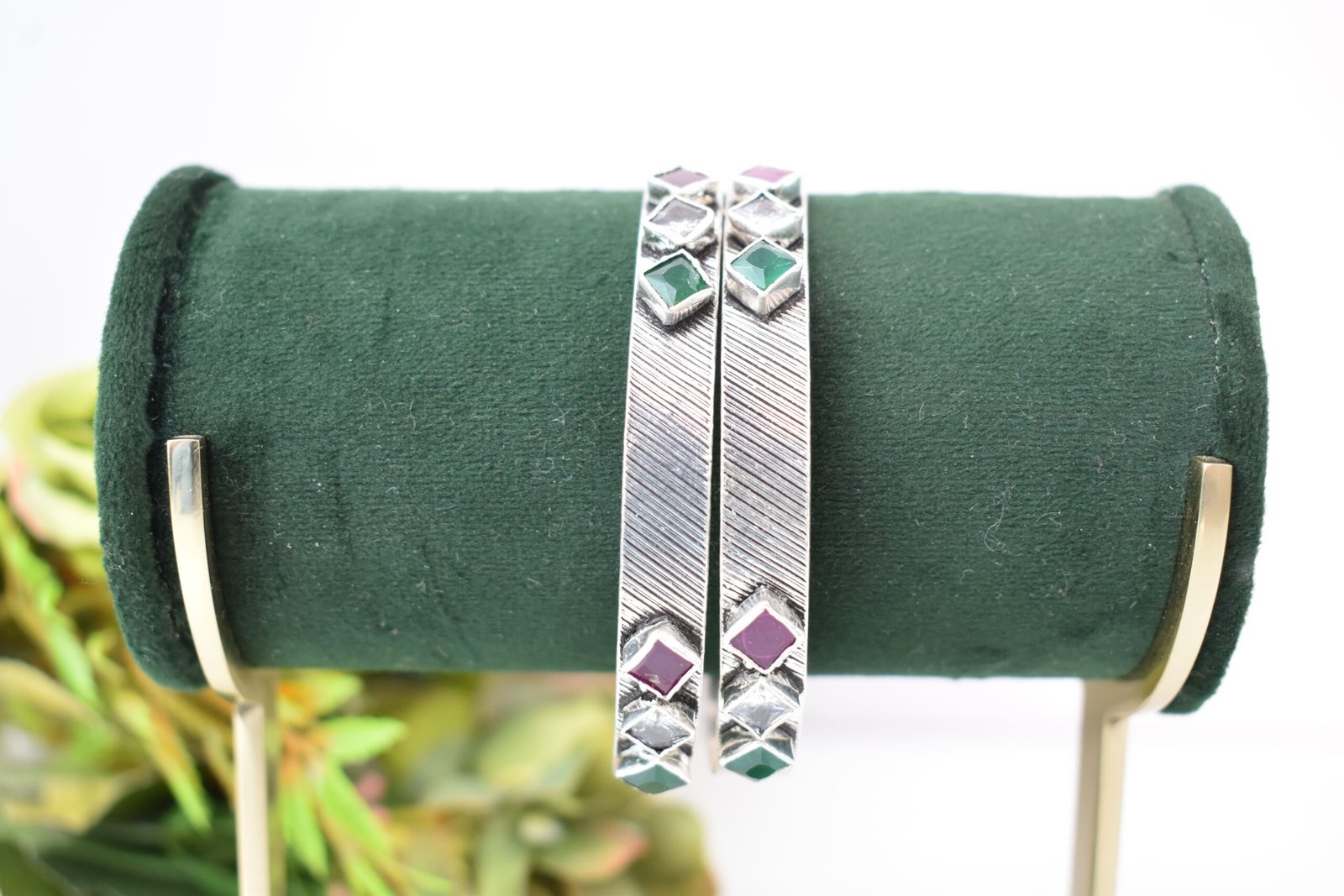 square-green-white-red-sequence-silver-kada2 Close-up of two 925 sterling silver kadas standing upright, featuring a diagonally textured silver band and a sequence of square-cut green, white, and red stones set along the edges against a soft floral background.