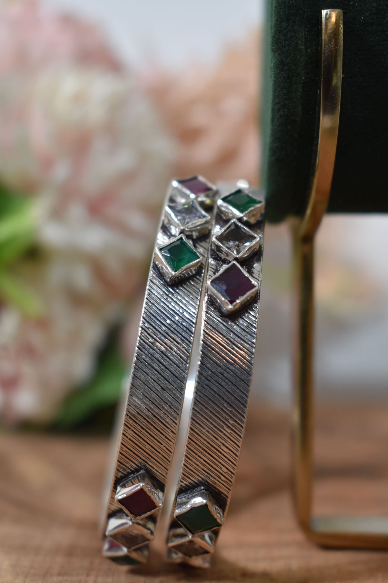 square-green-white-red-sequence-silver-kada3 Close-up of two 925 sterling silver kadas standing upright, featuring a diagonally textured silver band and a sequence of square-cut green, white, and red stones set along the edges against a soft floral background.