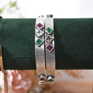 Close-up of two 925 sterling silver kadas standing upright, featuring a diagonally textured silver band and a sequence of square-cut green, white, and red stones set along the edges against a soft floral background.