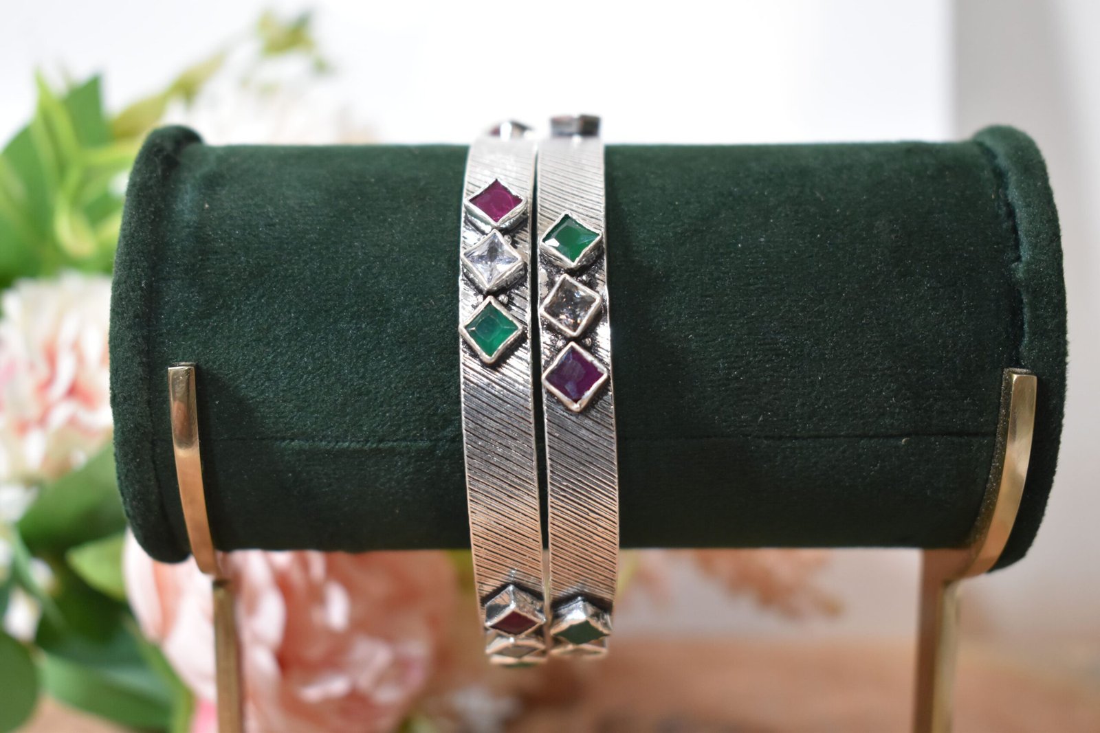 square-green-white-red-sequence-silver-kada4 Close-up of two 925 sterling silver kadas standing upright, featuring a diagonally textured silver band and a sequence of square-cut green, white, and red stones set along the edges against a soft floral background.