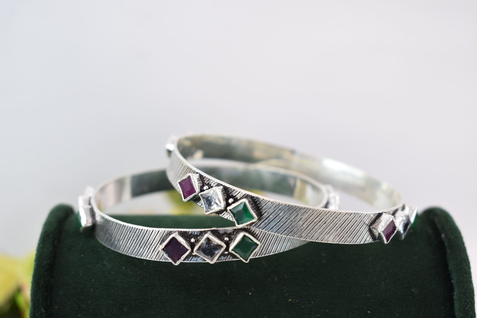 square-green-white-red-sequence-silver-kada5 Close-up of two 925 sterling silver kadas standing upright, featuring a diagonally textured silver band and a sequence of square-cut green, white, and red stones set along the edges against a soft floral background.