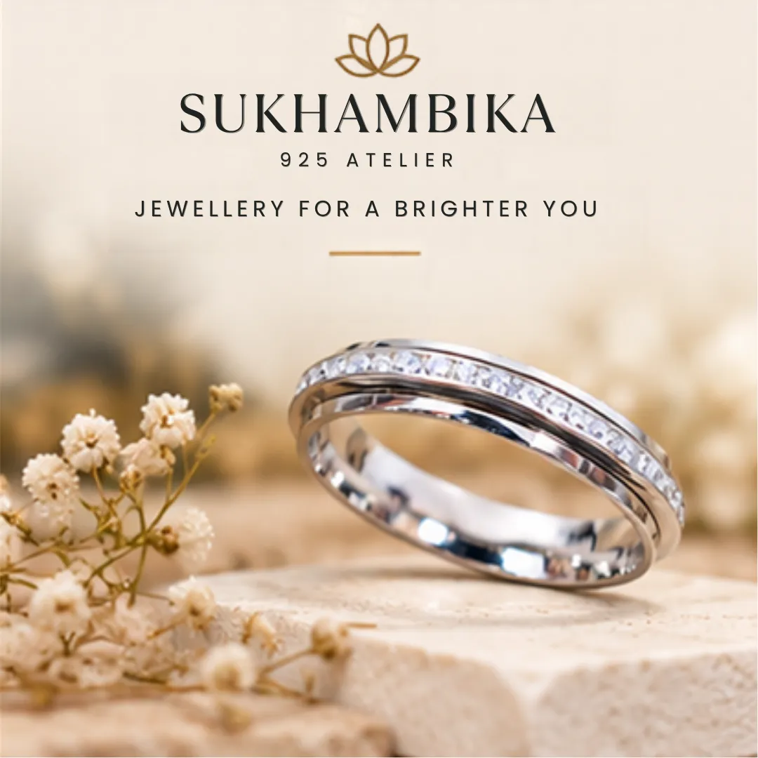 scintillating-cz-stone-finished-silver-band-sukhambika (9) Promotional product image of the scintillating cz stone finished silver band resting on a stone block with delicate white flowers, featuring the text "Sukhambika 925 Atelier Jewellery for a Brighter You".